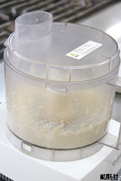 白い土台の上に置かれた透明な容器の中にベージュ色のペースト状の食材が入っており、上部に投入口がある透明な蓋が閉じられたフードプロセッサーの写真