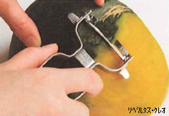 半分に切ったかぼちゃを安定させ、右手のピーラーを硬い外皮に押し当てて手前に引くことで、緑色の皮を薄く削り取っている下ごしらえの様子を捉えた写真