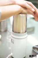 白い土台に細長い透明な容器がセットされ、中に白っぽい液体が入った状態で、動作させるために上部を両手で上からしっかりと押さえている、コンパクトな家庭用ミルミキサーを使用している場面の写真
