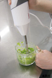 目盛り付きの透明な計量カップに入れられた鮮やかな緑色のペースト状の食材を、白い本体のハンディフードプロセッサーを片手で垂直に差し込み、もう一方の手でカップを固定しながら滑らかに攪拌している、調理作業中の様子を捉えた写真