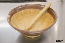 内側に細かな溝が刻まれた茶色の大きな陶磁器製のすり鉢と、その中に斜めに立て掛けられた木製の太いすりこぎ棒を写した写真