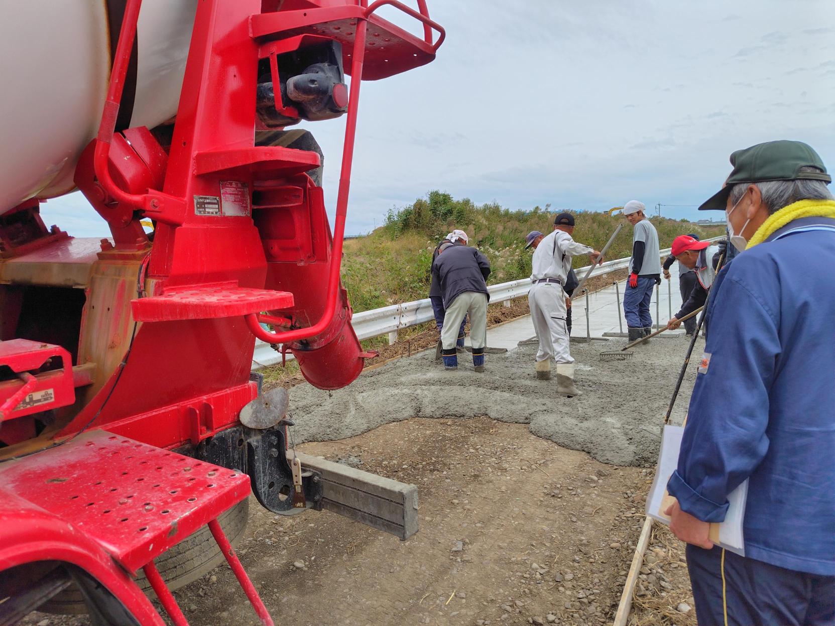 赤いコンクリートミキサー車の後部から排出された生コンクリートを、複数の作業員たちがスコップやトンボを使って平らに均している農道の舗装の様子を写した写真