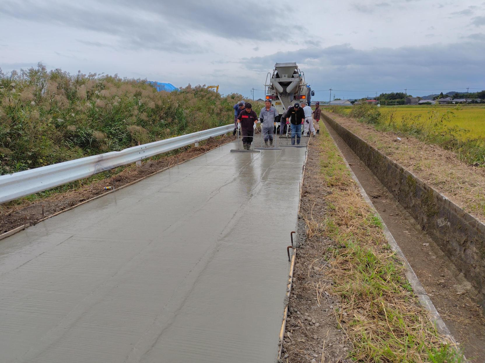 背後にはのどかな田畑が広がる田園地帯の農道において、ミキサー車から流し込まれた生コンクリートを作業員たちが長いトンボを使って平坦に仕上げて舗装工事が完了に近づく様子を捉えた写真