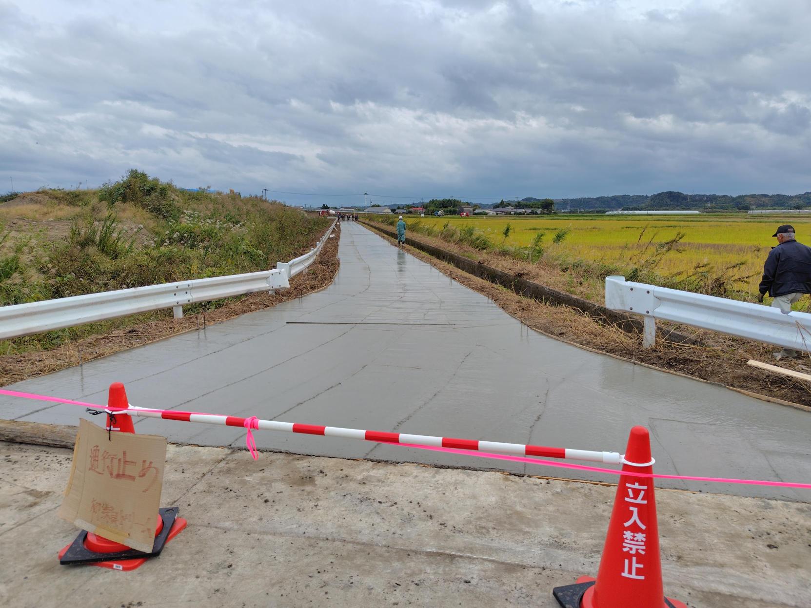 手前に「立入禁止」と書かれた赤いコーンとバーが設置され、その奥に新しく敷設されたばかりの滑らかなコンクリートの道が真っ直ぐ続き、右側に広がる黄金色の田んぼが広がっている農道の舗装工事現場を捉えた写真