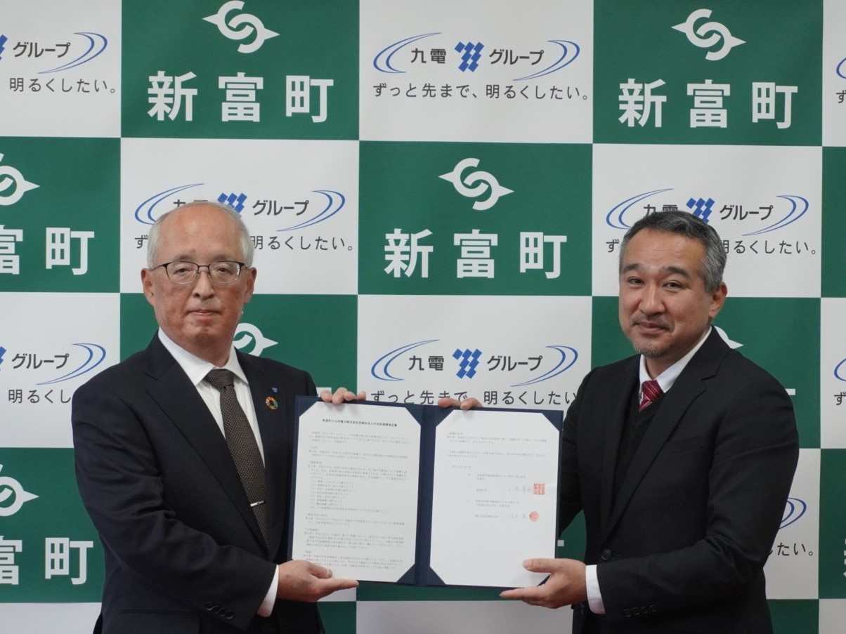 「新富町」の文字と市章、及び「九電グループ」のロゴが並ぶ背景の前で、スーツ姿の久富木支店長と小嶋町長が一緒に見開きの協定書を掲げ、カメラに向かって穏やかな表情で並んでいる写真