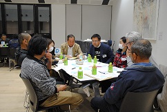 明るい会議室で「D」と書かれた札のあるテーブルを囲み、参加者の男女数名が、広げられた資料を真剣な表情で見つめながら活発に意見を交わしている、ワークショップの様子を捉えた写真