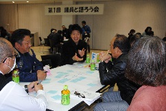 付箋が貼られたワークシートを囲み、身振り手振りを交えて笑顔で語り合う男性と、その話を真剣に聞く参加者たちの活気あるグループワークの様子を写した写真