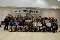 「第1回 議員と語ろう会」の横断幕が掲げられた会場で、最前列に並んだ椅子に座る人々とその後ろに立つ人々が、室内で集合写真を撮影している記念写真