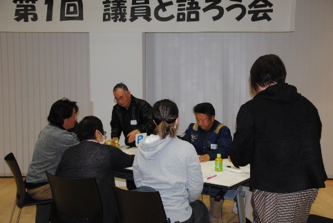 「第1回 議員と語ろう会」と書かれた横断幕の下、会議室のテーブルを囲んで数人の参加者が座り、資料や飲み物を前にして真剣な表情で対話や作業を行っている様子を捉えた写真