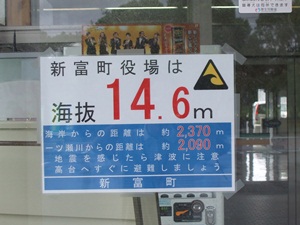 新富町役場の海抜が14.6メートルであることを大きな赤字で示し、海岸や一ツ瀬川からの距離、地震発生時の津波への注意喚起と避難を促す文言が記載された、屋外に掲示されている津波避難計画の案内板の写真