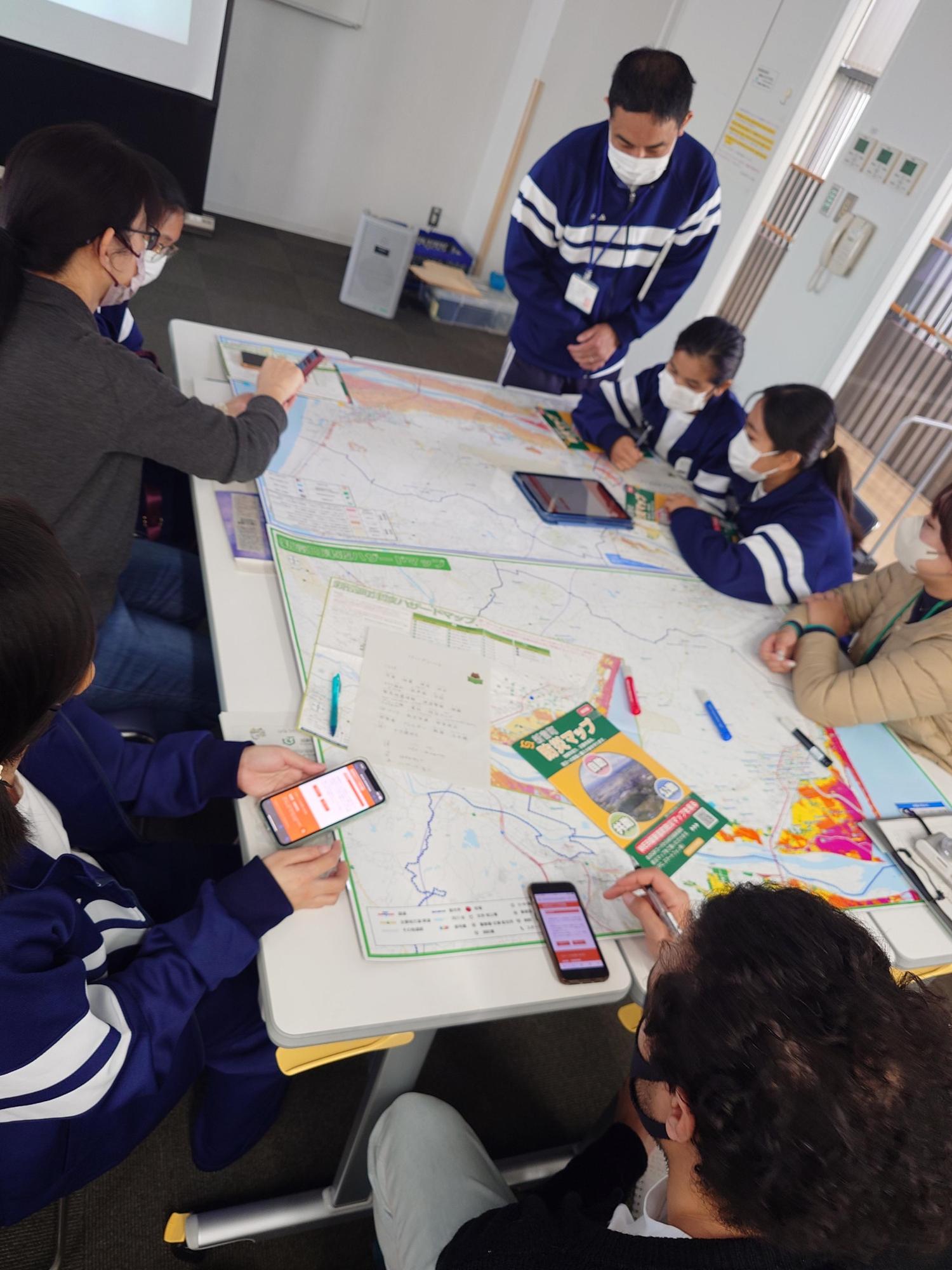 テーブルに広げられた大きな防災ハザードマップを囲み、スマートフォンやタブレットを手に持ちながら、マスクを着用した複数人の参加者が真剣な表情で地域の危険箇所や避難経路を確認し合っている防災教室の様子の写真