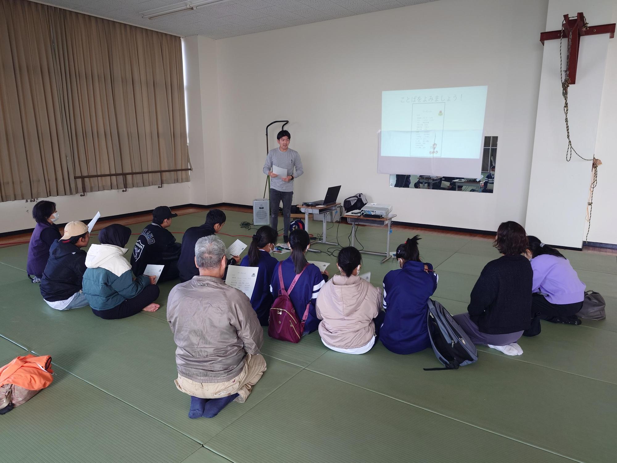 プロジェクターで投影されたスライドを前に、男性講師が資料を手に立ち、畳の部屋に座って熱心に説明を聞きながら手元の資料を確認している交通ルール教室の様子を捉えた写真