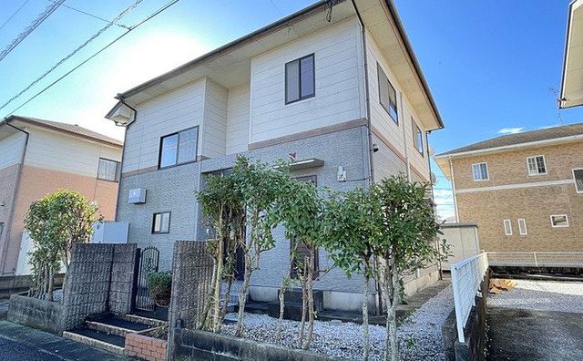 ベージュとグレーのツートンカラーの二階建て住宅で、手前には数本の低い庭木が植えられ、左側には石造りの門柱と黒いアイアンゲートがあり、建物の周囲には砂利が敷き詰められている一戸建住宅の物件の写真