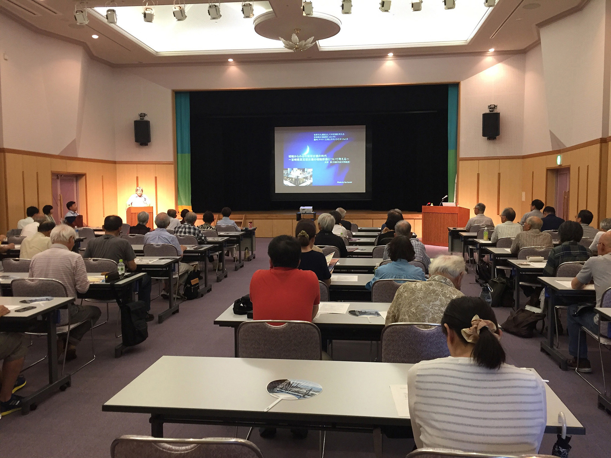 ホールのステージ上で、前方スクリーンに画像が大きく映し出され、演壇に立つ講師の話を多くの参加者たちが静かに聞き入っている講演会の様子を捉えた写真