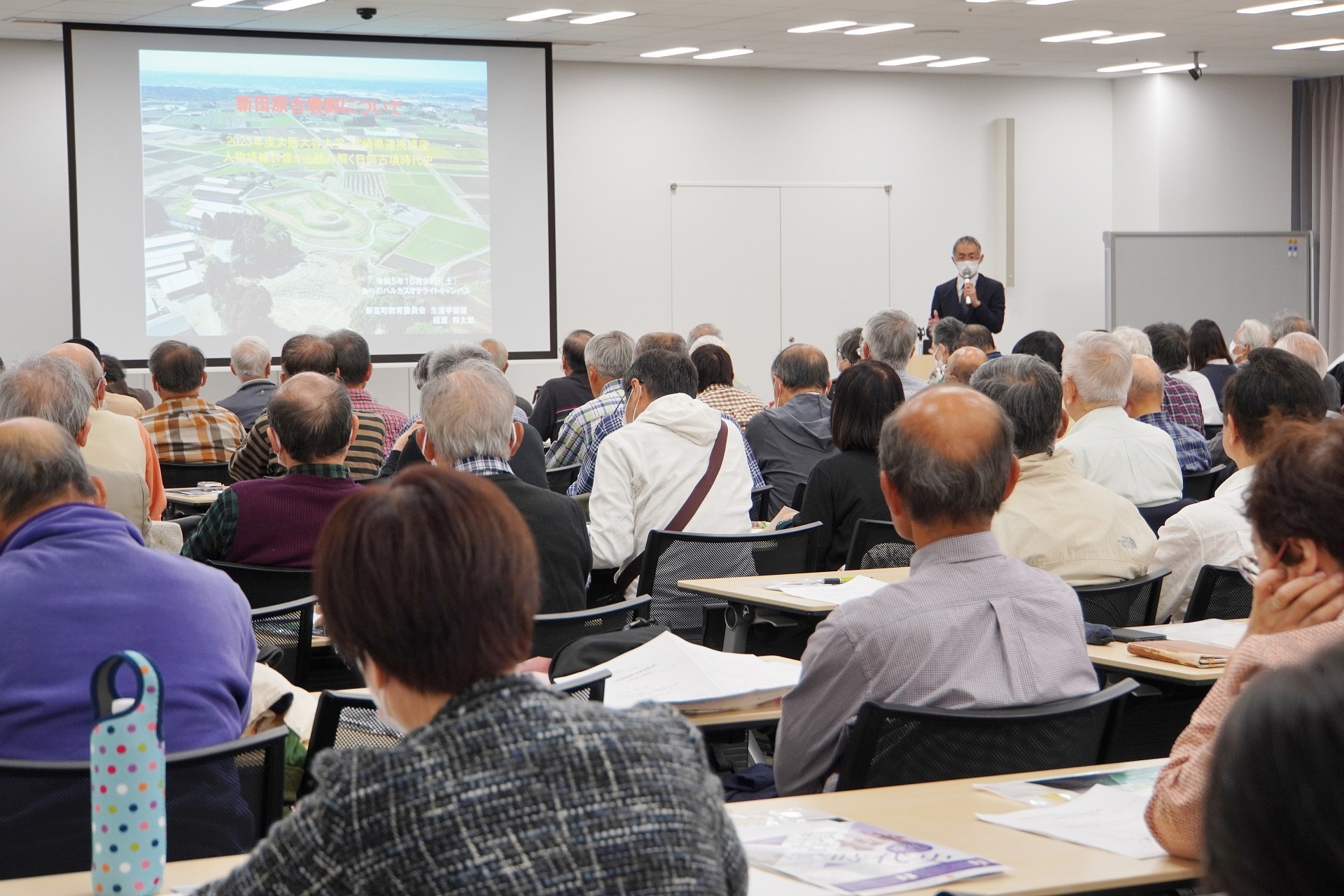 会議室の前方スクリーン資料の写真が大きく映し出され、スーツ姿の講師がマイクを手に説明を行う中、会場を埋め尽くした多くの参加者たちが手元の資料を見ながら静かに聞き入っている真大阪大谷大学講座の様子を捉えた写真