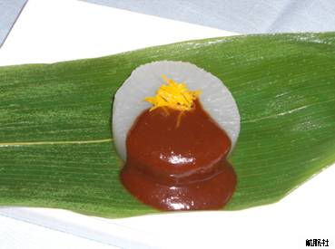 笹の葉の上に載せられた白い円形の大根にたっぷりと赤味噌がかけられ、細長く刻まれた黄色い柚子の皮が美しく添えられている、大根の味噌かけ柚子和えの写真