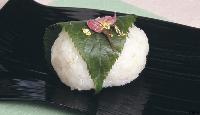 緑色の桜の葉で包まれた上に桜の花びらの塩漬けと豪華な金箔が散らされたつぶつぶとした米の質感が残る白い俵型の桜もちの写真