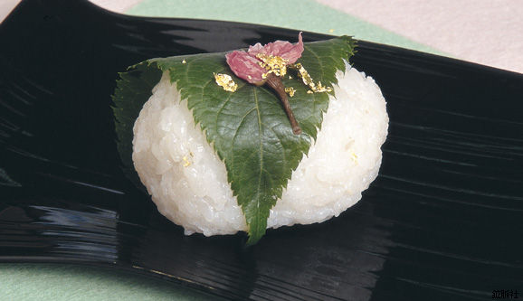 緑色の桜の葉で包まれた上に桜の花びらの塩漬けと豪華な金箔が散らされたつぶつぶとした米の質感が残る白い俵型の桜もちの写真