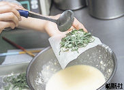 千切りした青じそをクッキングペーパーの上において手に持ち、衣をお玉でかけている写真