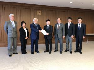 スーツ姿の男女7名が横一列に並び、中央の2名が要望書を手に持ち受け渡しをしている様子の写真
