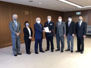 マスクを着用した男女7名が横一列に並び、中央で紺色のスーツを着た男性と制服を着た男性が要望書を持っている写真