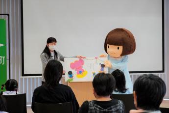 マスクをした女性と、茶色のボブヘアのおとみちゃんが、ピンク色のゾウが描かれた大きな絵本を広げて子供たちに読み聞かせをしている様子の写真