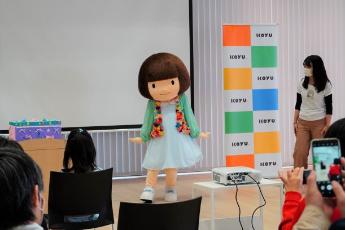 茶色のボブヘアのおとみちゃんが、カラフルな飾りの付いたカーディガンを羽織ってステージであいさつをする様子の写真