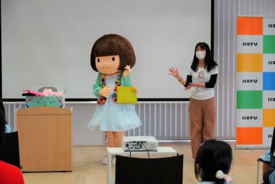 ボブヘアのおとみちゃんが黄色いバッグを手に持ち、隣でマスク姿の女性がポーズを交えて紹介している様子の写真