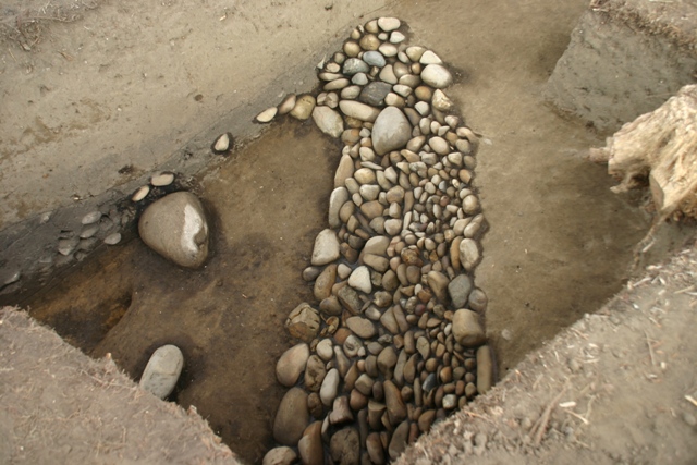 土を四角く掘り下げた発掘調査のトレンチ内部において、大きさの異なる丸みを帯びた河原石のような石が縦長に密集して敷き詰められている様子を斜め上から近距離で撮影した、前方部から後円部とがつながる部分「クビレ部」を写した写真