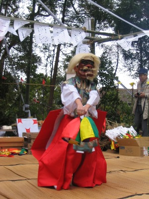 屋外に設営された舞台の上で、白髪と恐ろしい形相の面をつけ、鮮やかな赤い袴を纏った人物が、黄色・緑・赤の三色の飾りが付いた道具を手に持って舞を披露している写真