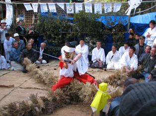 多くの見物人に囲まれた屋外の祭事場にて、白い装束と赤い袴を身に纏い、白い布で鉢巻をした舞い手が、蛇にみたてた太い注連縄の上で真剣を構えて精神を集中させている、緊迫感あふれる勇壮な蛇切りを捉えた写真