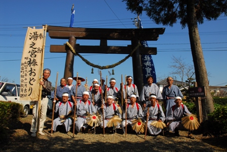 青空の下、左端には「奉納 湯之宮棒おどり」と記された幟が掲げられ、神社の鳥居の前に伝統的な祭礼衣装を身にまとった保存会の男性たちが勢揃いしている写真