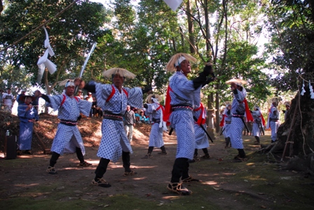 木々に囲まれた神社内の広場で、編み笠を被り白と紺の紋様の着物に赤い襷を掛けた複数の男性たちが、剣を手に持ち力強く演舞を披露している、伝統芸能「棒おどり」の躍動感あふれる奉納儀式の写真