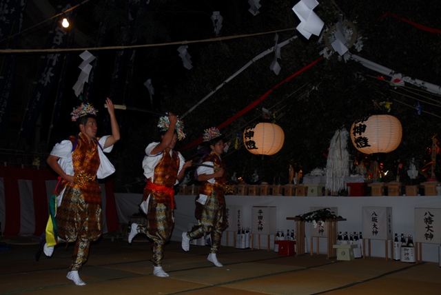 夜の静寂に包まれた屋外の特設舞台において、華やかな花飾りを頭に乗せ、金色の紋様が施された豪華な衣装を纏った三人の若者が、手に刀を持ちながら一斉に力強く躍動する、伝統的な夜神楽の奉納演舞を捉えた写真