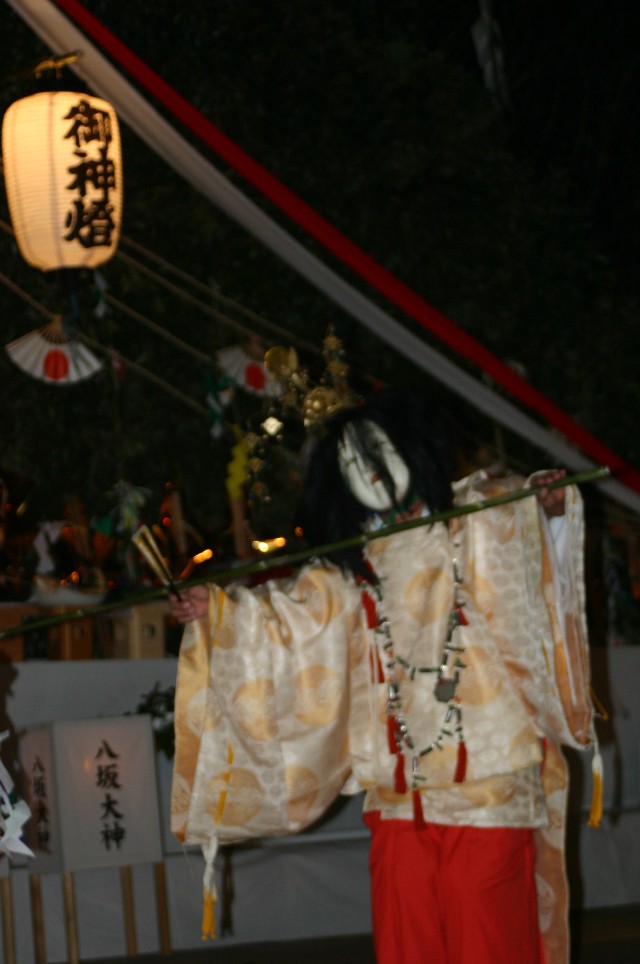 夜の静かな神楽舞台において、白く穏やかな表情の面と黒い長髪をつけ、金色の丸紋が散りばめられた豪華な白い装束に赤い袴を纏った舞い手が、一本の長い竹の棒を水平に保持しながら舞を披露する神聖な夜神楽のワンシーンの写真