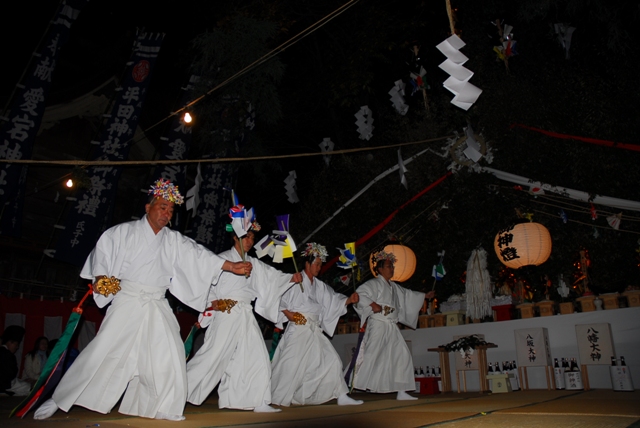 夜の神楽舞台において、頭に鮮やかな花飾りを載せ、清らかな白い装束と袴を身にまとった四人の舞い手が、右手に金色の鈴を持ち、左手で色とりどりの飾りが付いた道具を掲げながら一列に並んで舞う、厳かで活気ある奉納行事のワンシーンの写真