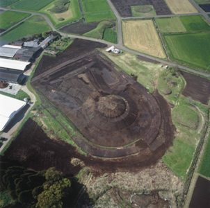 平坦に整地された広大な黒い土の敷地中央に、円形に盛り土がなされた前方後円墳のような遺跡が保存されており、その周囲を緑の畑や農道、白い屋根の建物が囲んでいる様子を上空から写した写真