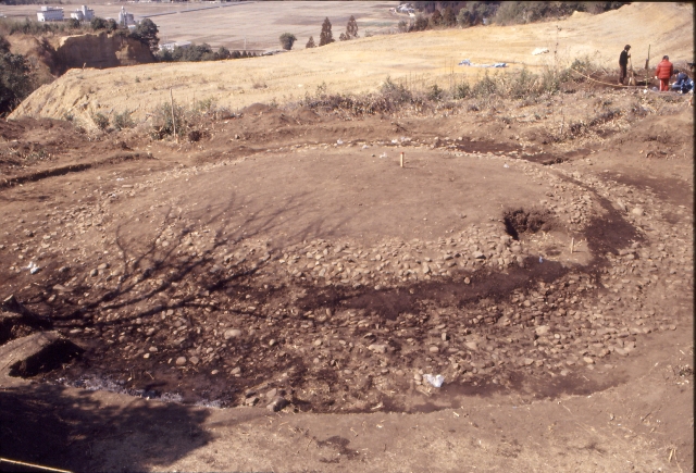 見晴らしの良い高台にある発掘調査現場にて、円形状に石が敷き詰められた配石遺構が土の中から露出しており、周囲では作業員が調査を行う様子や遠方に広がる平野部までを一望できる様子を写した写真