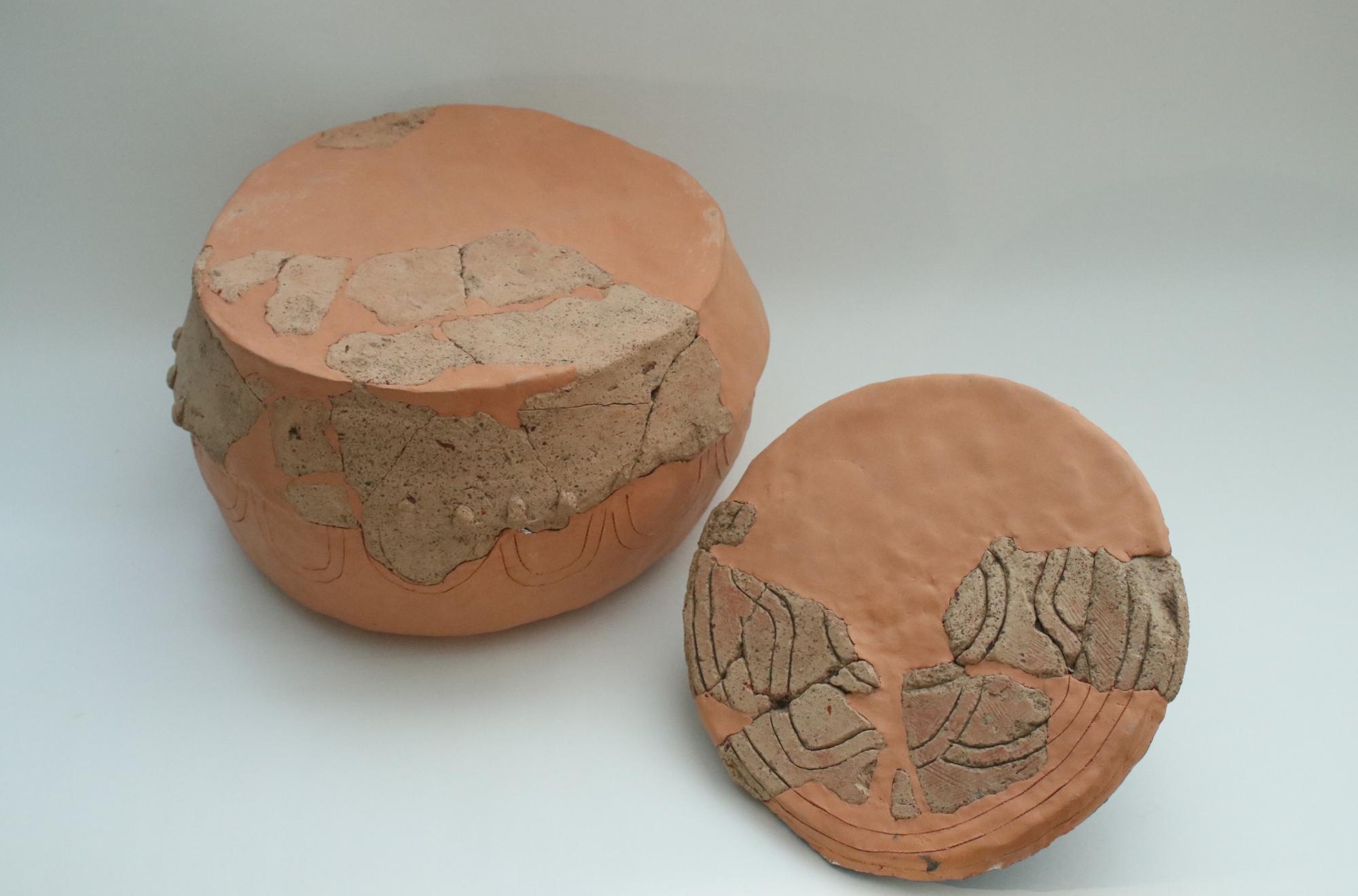 丸みを帯びた器の胴体部分、右側には円盤状の蓋のようなパーツが置かれ、表面には波状の文様や細かな凹凸が施され修復された赤茶色の素焼きの太鼓形埴輪の写真