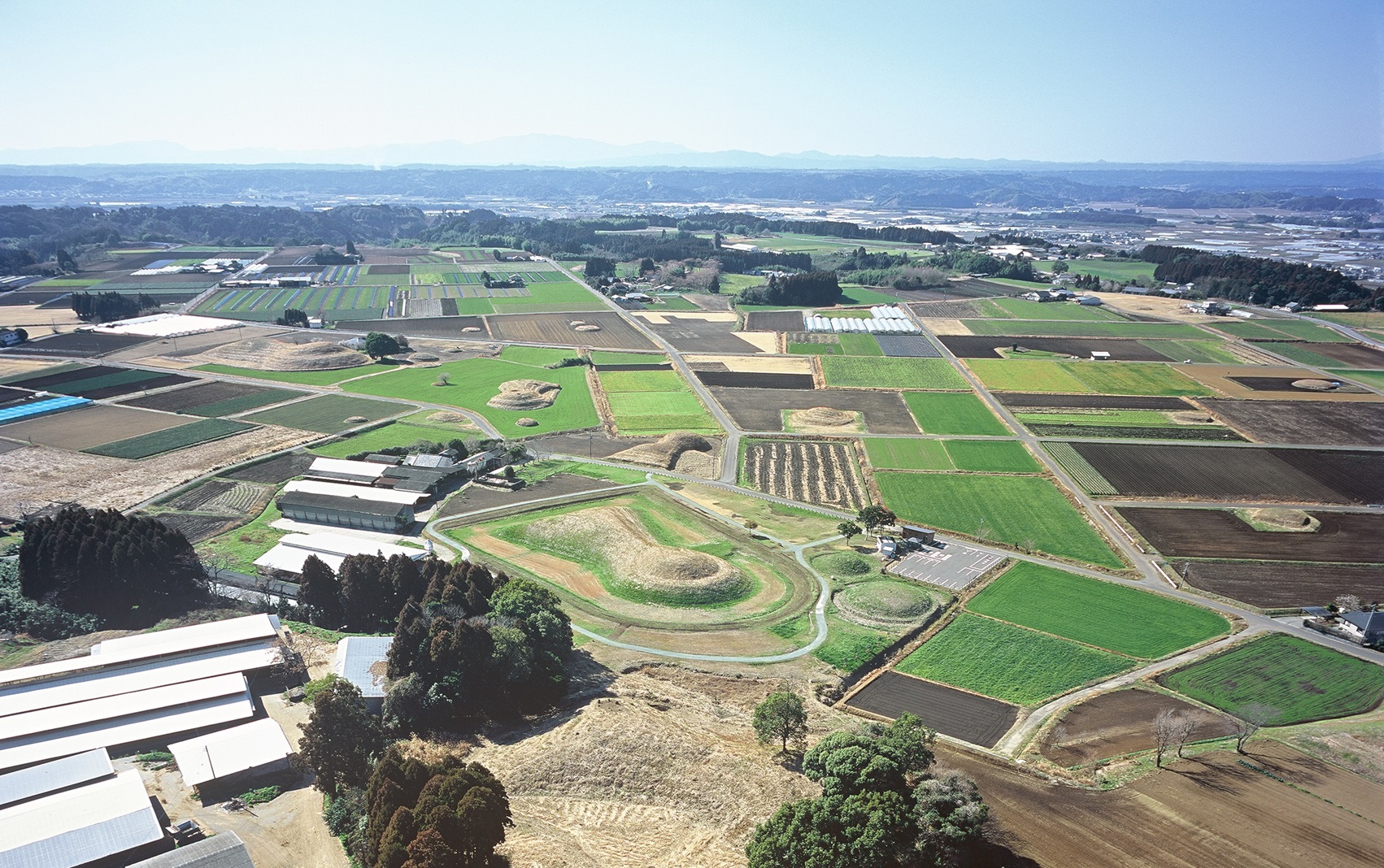 広大な田畑が整然と区画された平野の中に、鍵穴のような形をした前方後円墳や円墳が点在しており、手前には白い屋根の建物や木々、遠くには霞んだ山々が広がる、歴史的遺構と農地が共存する風景を俯瞰で捉えた写真
