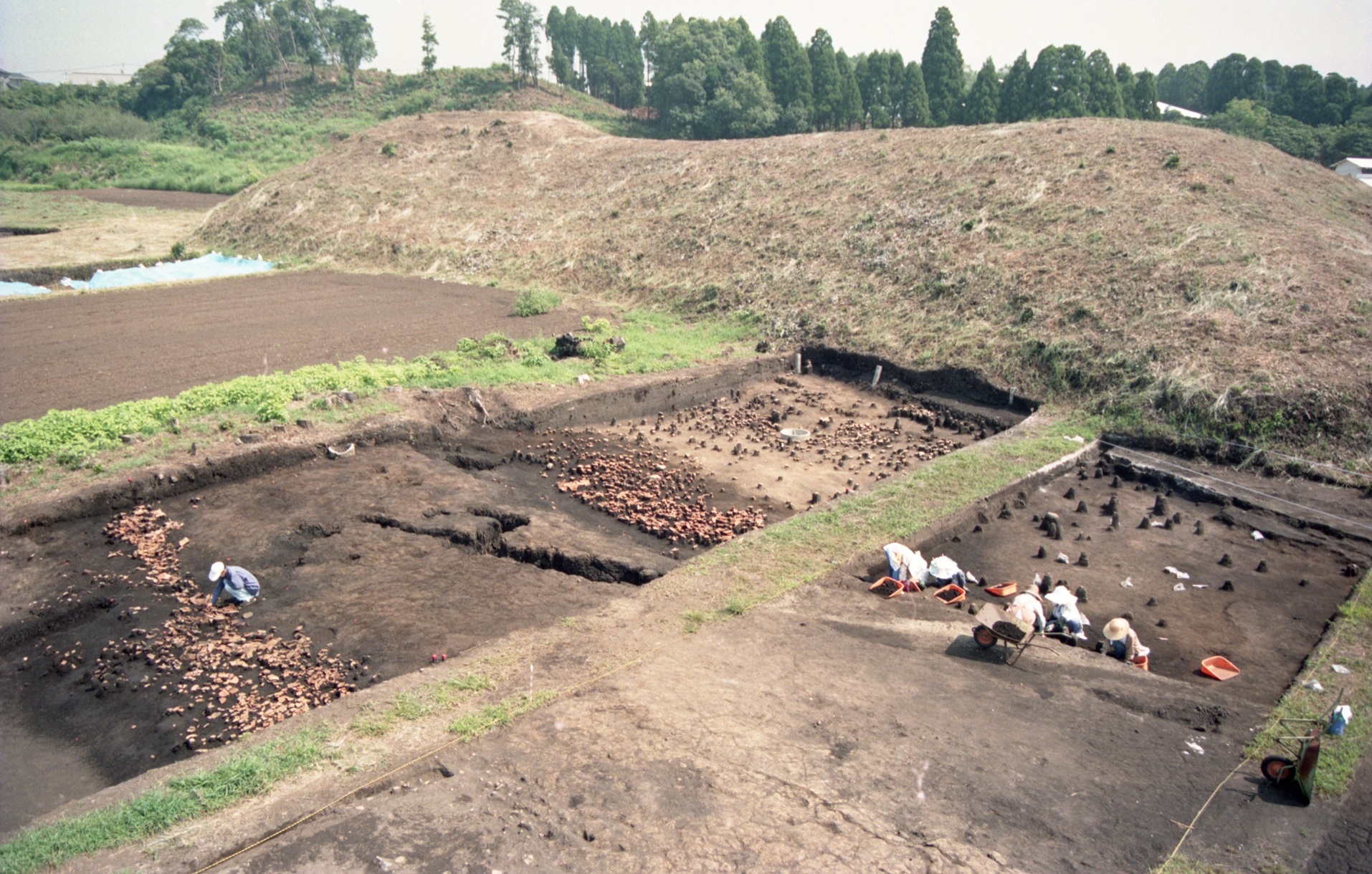 緩やかな丘のような古墳の麓に、長方形に区画された発掘調査現場が広がっており、作業服を着た数名の調査員が地面に膝をついて土器や遺構を慎重に掘り出している調査の様子を捉えた写真