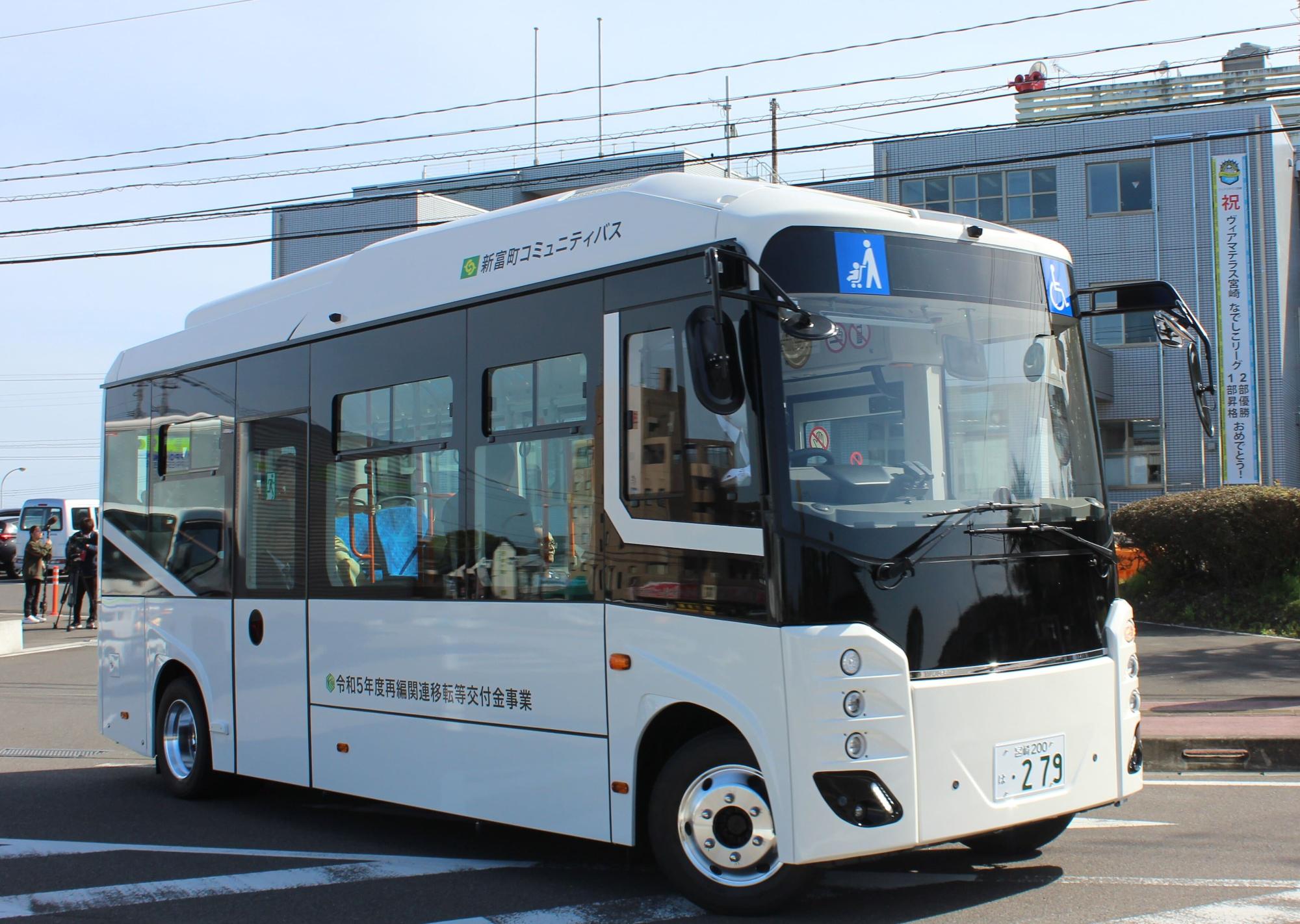 白を基調とした車体に「新富町コミュニティバス」と表記された小型のノンステップバスが道路を走行しており、車両にはベビーカーマーク、車椅子マークが掲示されているバリアフリー対応の新富町コミュニティバスの写真