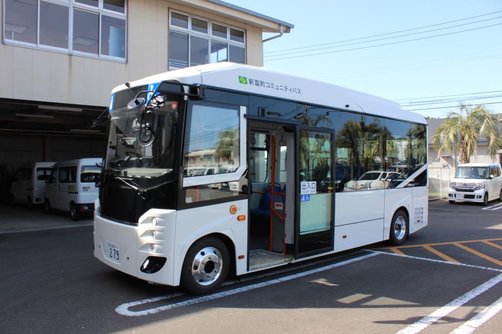 白を基調とした車体に「新富町コミュニティバス」のロゴが入り、乗降口のドアが開いた状態で駐車場に停車している小型のノンステップバスを、斜め前方から捉えた写真