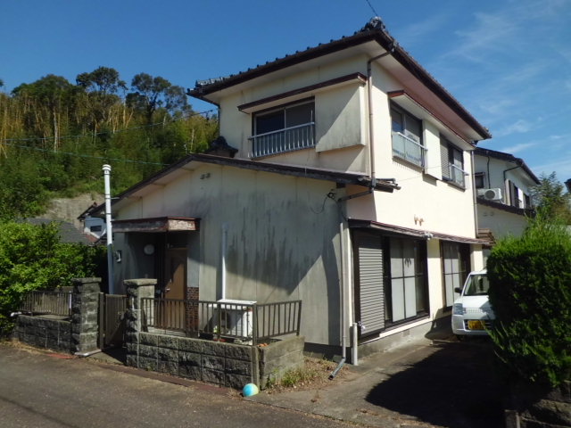 後方に緑豊かな山が迫る場所にある白壁の二階建て住宅で、手前には石積みの門柱がある玄関アプローチと右側に駐車スペースある一戸建て住宅の物件の写真